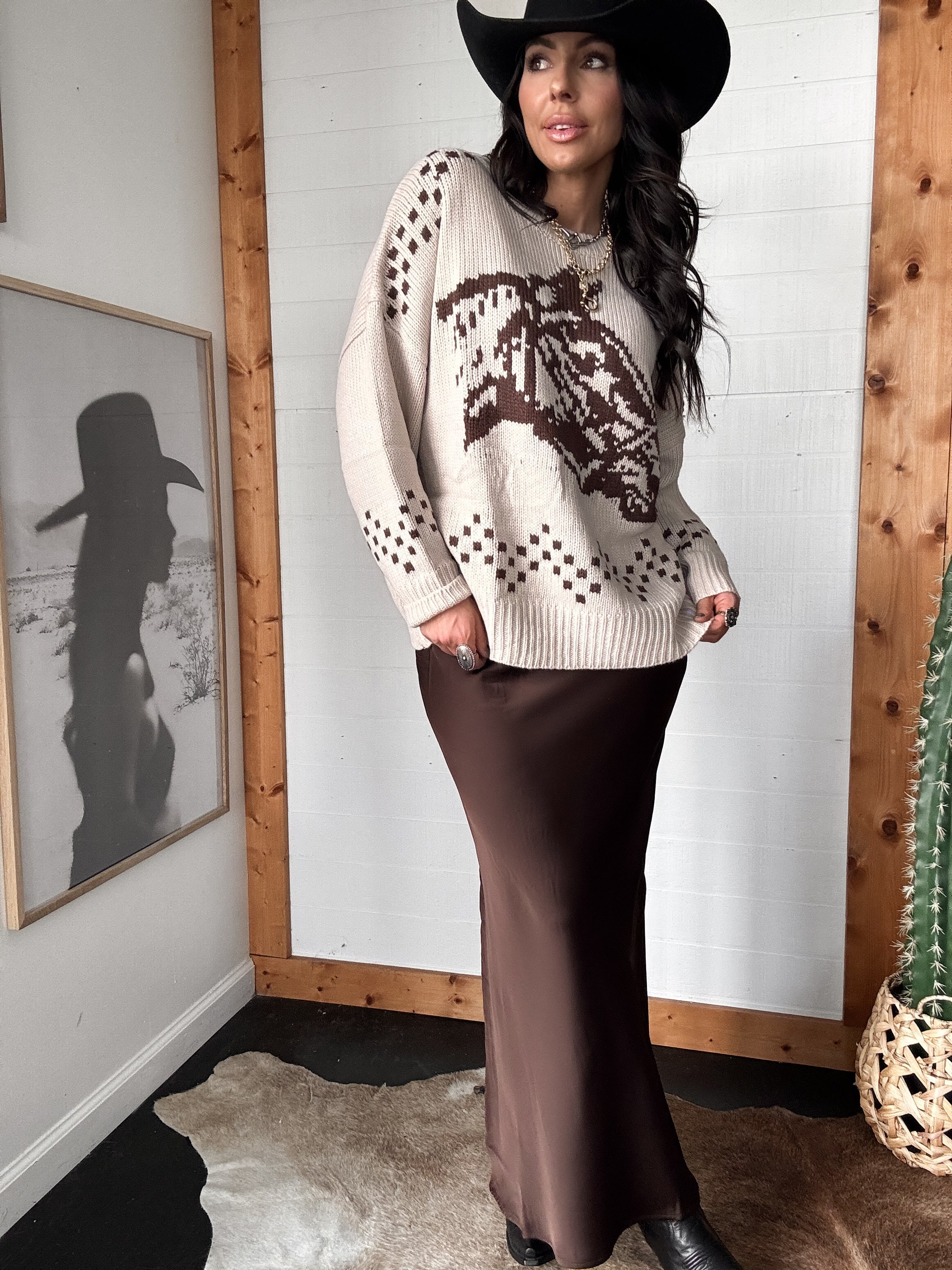 Woman in espresso satin maxi skirt, western graphic sweater, and black cowboy hat, standing indoors.