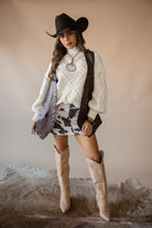 Woman in western boho outfit with black cowboy hat, cream sweater, cow print skirt, tan boots.