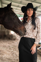 Woman in western boho outfit and black hat standing beside a brown horse in a barn setting