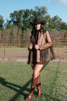 Woman in brown fringe western jacket, cowboy hat, and tall boots standing outdoors