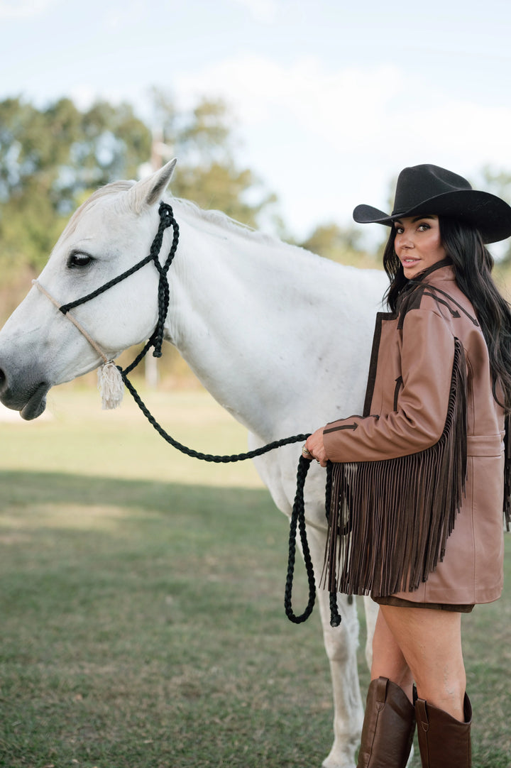 Woman in fringe western jacket, cowboy hat, and boots holding white horse outdoors