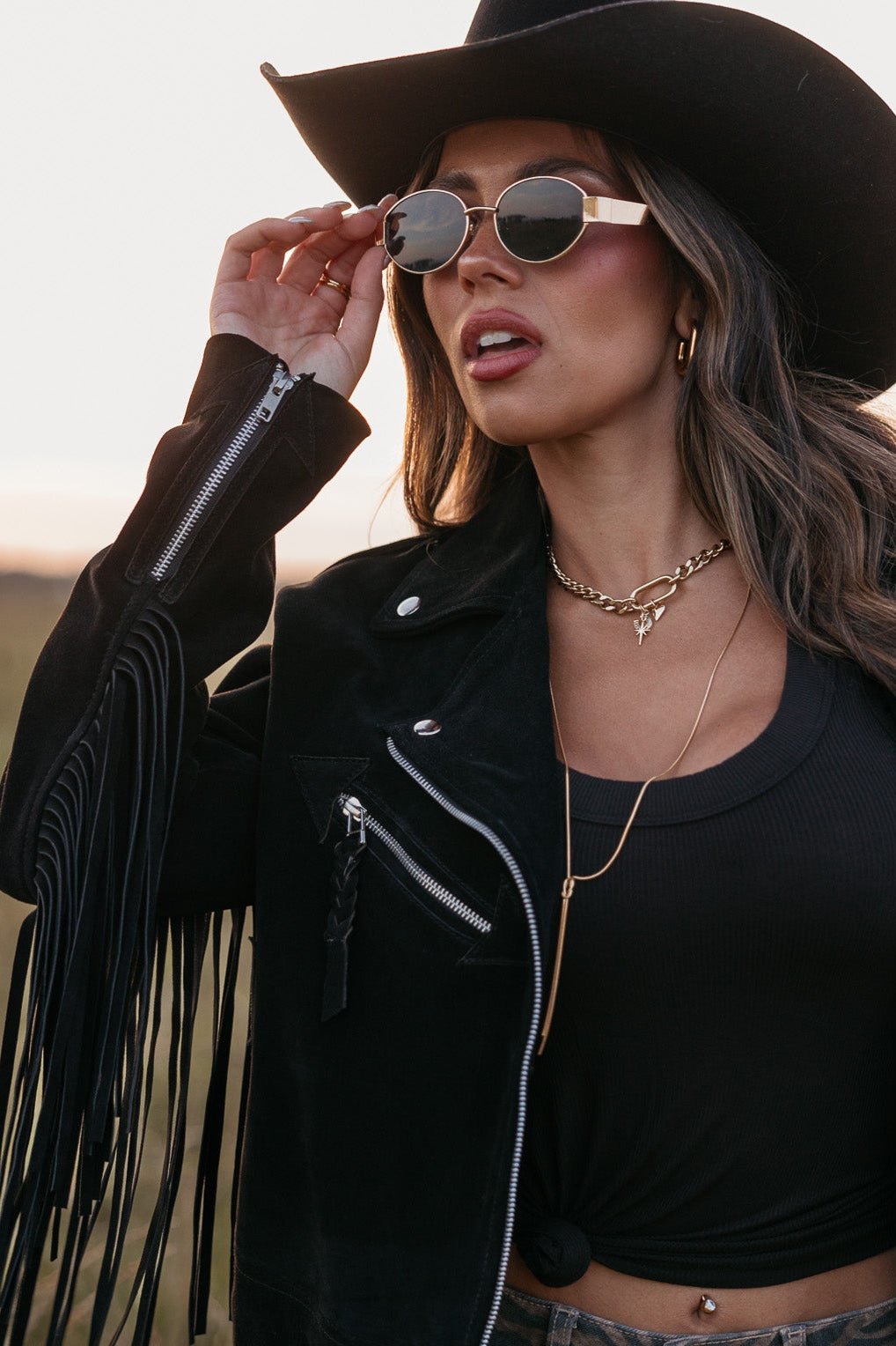 Woman in a black fringe jacket, black tank top, and gold oval framed glasses standing in a field.
