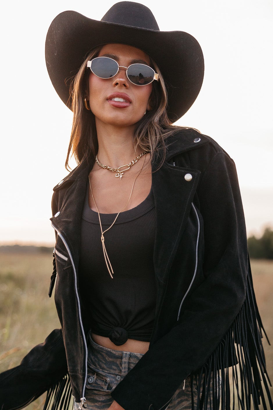 Woman in a black fringe jacket, black tank top, and gold oval framed glasses standing in a field.