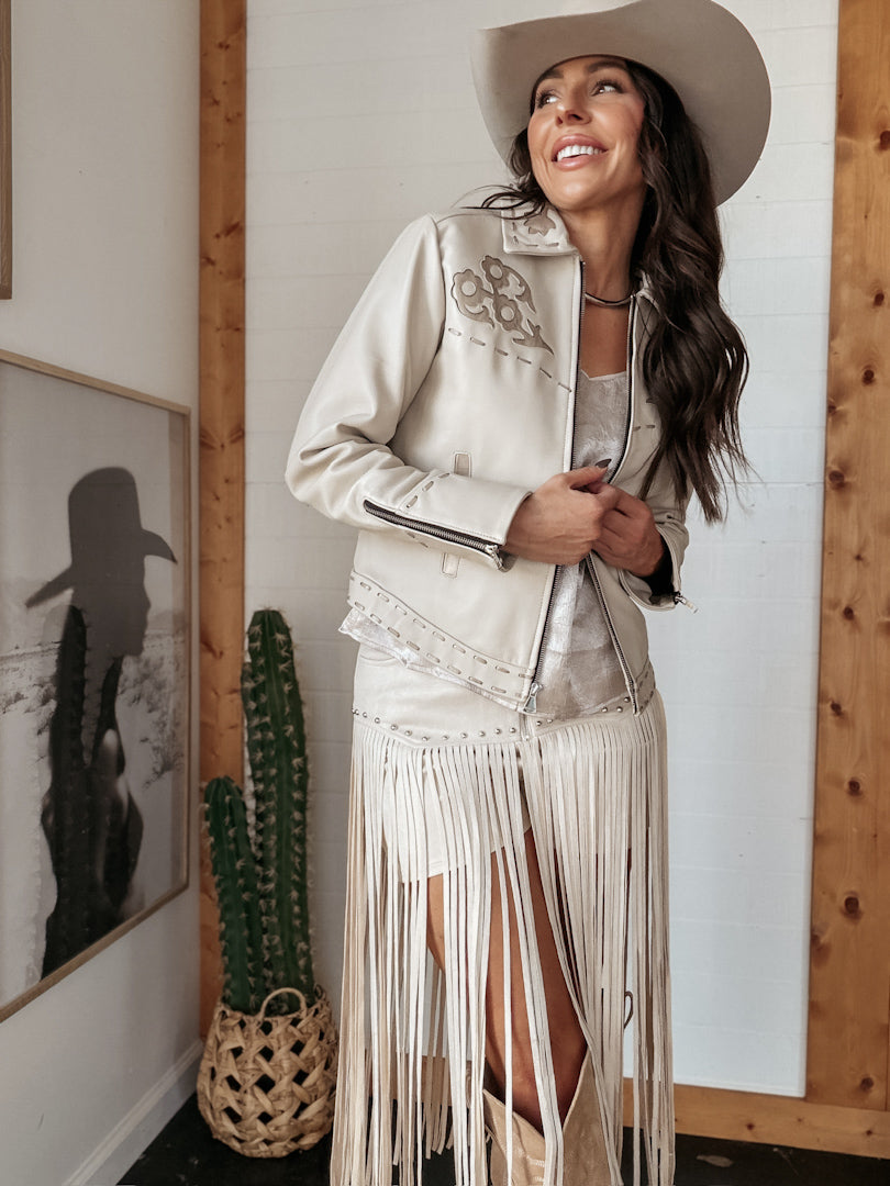 Woman in western boho fringe jacket, cream skirt, and cowboy hat, standing indoors.