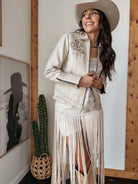 Woman in western boho fringe jacket, cream skirt, and cowboy hat, standing indoors.