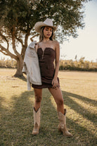Woman in strapless brown western dress, cowboy boots, and hat holding embroidered jacket outdoors