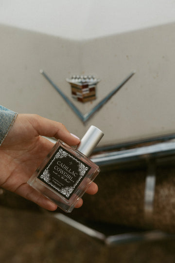 Perfume bottle labeled 'Cadillac Cowgirl' in front of a Cadillac emblem.