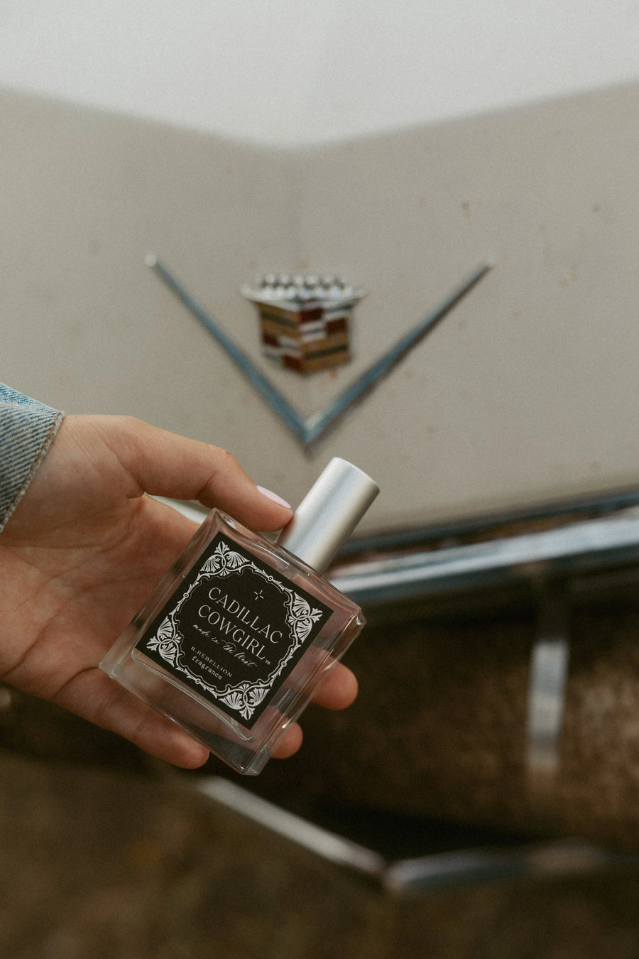 Perfume bottle labeled 'Cadillac Cowgirl' in front of a Cadillac emblem.
