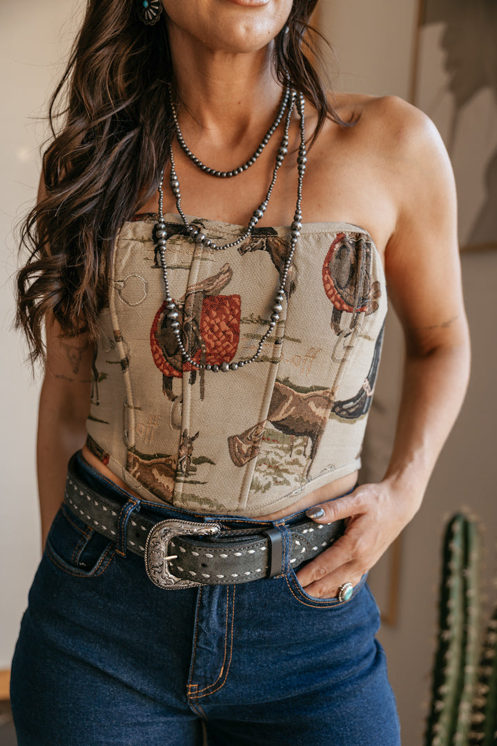 Model wearing an equestrian jacquard corset top with woven horse and saddle tapestry, styled with high-rise western denim, statement belt, and layered necklaces for an arena to after cowgirl outfit.