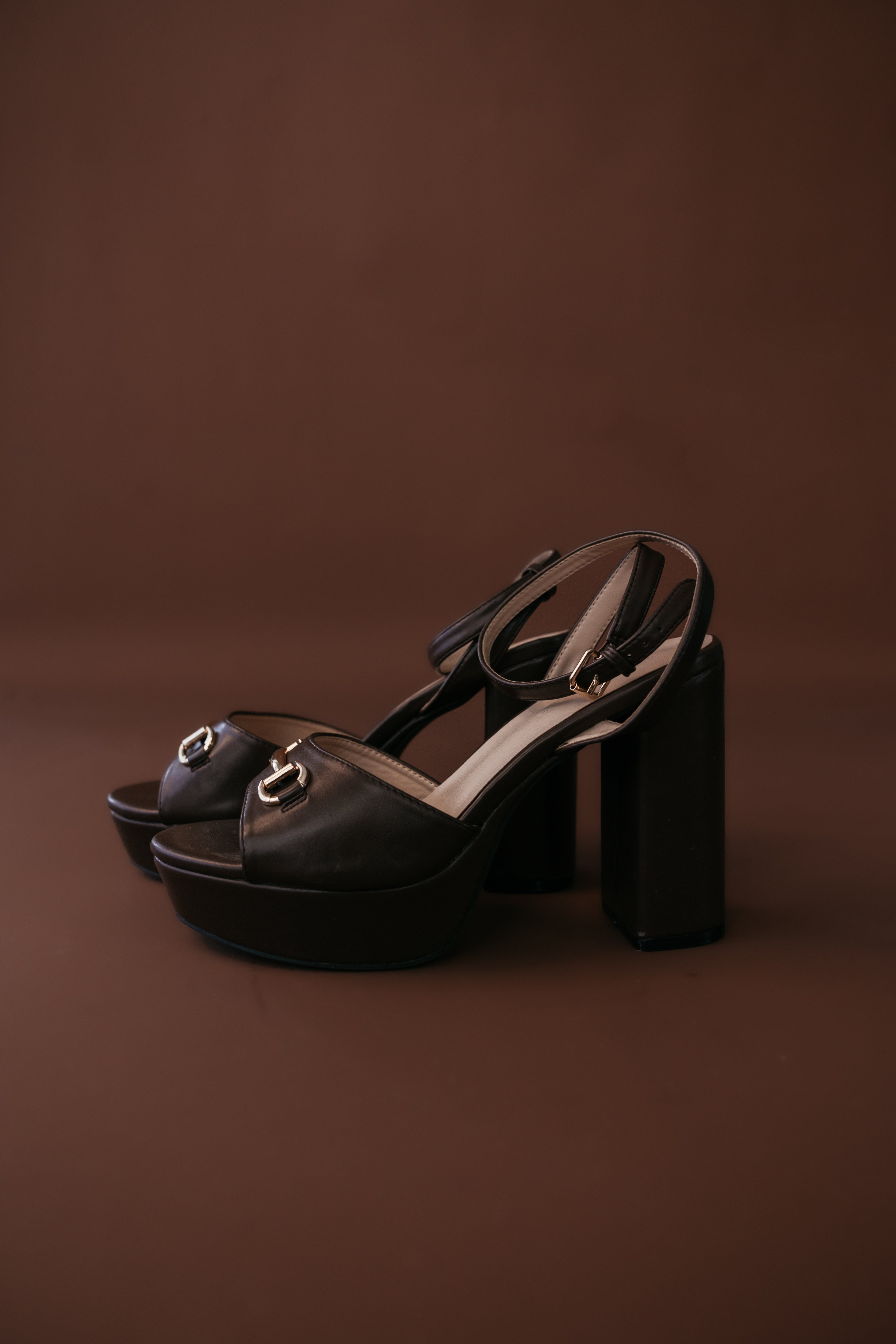 Black platform block heel sandals with gold horsebit detail on brown background