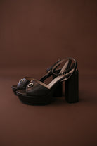 Black platform block heel sandals with gold horsebit detail on brown background