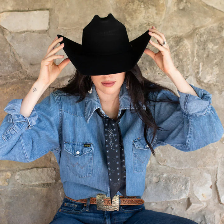 Person wearing a black cowboy hat and denim jacket against a stone wall.